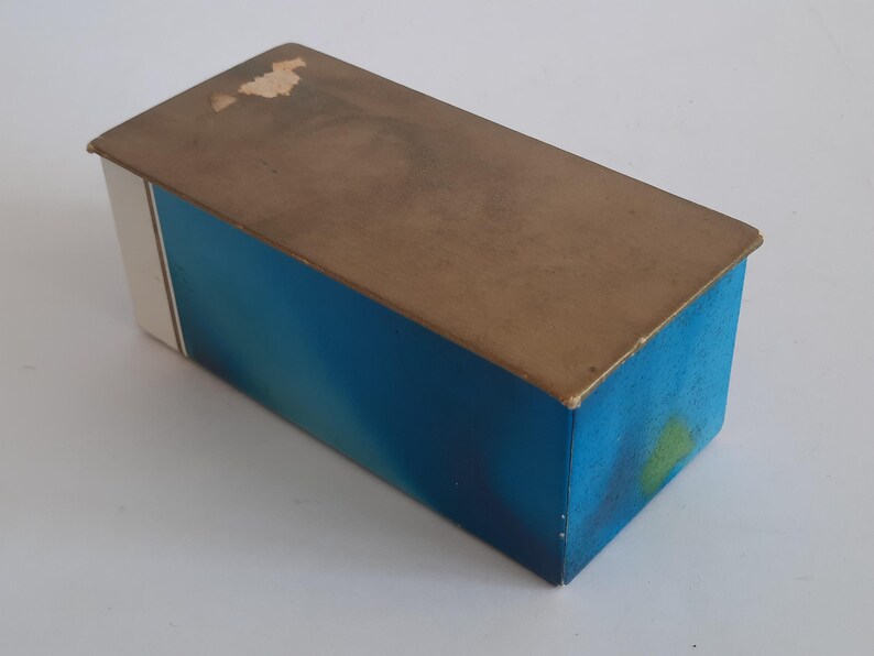 Puede incluir: Caja rectangular con un dise&ntilde;o degradado azul en los lados y una tapa marr&oacute;n. La caja tiene una franja blanca en un lado. La tapa muestra signos de desgaste. La caja es probablemente vintage o antigua.