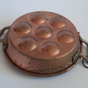May include: A copper pan with six round indentations and two brass handles. The pan has a decorative border around the edge.
