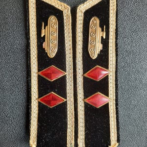 Puede incluir: Un par de hombreras militares negras con ribetes dorados e insignias rojas en forma de diamante. Un emblema de tanque dorado está en cada hombrera.