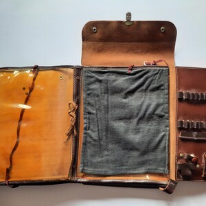 Puede incluir: Portafolio de cuero marrón abierto con interior beige claro y bolsillo de plástico transparente. El portafolio tiene un cierre de metal y múltiples compartimentos para bolígrafos y otros artículos. Tiene varios bolsillos para guardar.