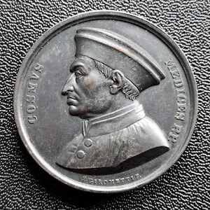 Puede incluir: Medalla redonda de color bronce que presenta el perfil de un hombre con sombrero y vestimenta abotonada. La inscripción "COSMVS" es visible a la izquierda y "MEDICES P.P." a la derecha. El nombre del artista, "P. GIROMETTI", está en la parte inferior.