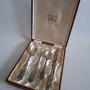 Op de afbeelding: Een vierdelige zilverkleurige bestekset bestaande uit een lepel, vork, kleine lepel en mes, elk met gedetailleerde reliëfpatronen op de handvatten. De set wordt gepresenteerd in een donkerrode doos met een crèmekleurige satijnen voering en Berndorf-logo.