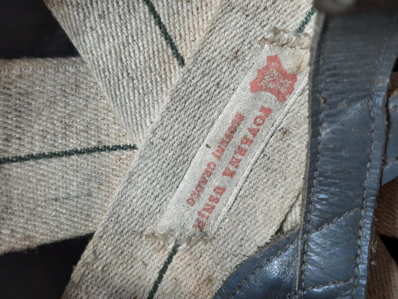 K&ouml;nnte beinhalten: Ein beigefarbener Canvas-Riemen mit einem d&uuml;nnen gr&uuml;nen vertikalen Streifen und einem wei&szlig;en Stoffetikett mit rotem Text, der TOVARNA USNJI SOGNI GRADEC lautet. Der Riemen ist an einem dunkelbraunen Lederteil mit sichtbaren N&auml;hten am Rand befestigt.