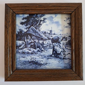 May include: A square ceramic tile featuring a blue and white monochromatic illustration of sheep shearing in a rural landscape with a thatched barn and windmill, encased in a dark brown wooden frame with a beveled edge and visible wood grain.