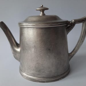 Puede incluir: Una tetera de color plateado con tapa y asa. La tetera tiene un pico y una base redondeada. El asa es curva y está unida al lateral de la olla. La tapa tiene un pequeño pomo en la parte superior. La tetera es de metal y tiene un acabado cepillado.