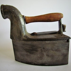 May include: A vintage cast iron flat iron with a wooden handle. The iron has a long spout on the side and is in a worn, aged condition.
