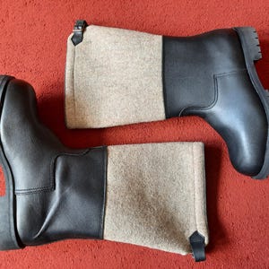 May include: A pair of black leather boots with a beige felt upper section. The boots have a rugged, ridged sole and are set against a red background. The boots are designed for cold weather.
