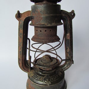 Könnte beinhalten: Eine antike, rostige grüne Metall-Petroleumlampe mit einem Glaszylinder und einem Metallgriff. Die Lampe hat einen federbelasteten Dochtregler und eine Metallkappe mit dem Text "Feuer Hand" darauf.