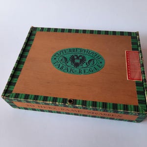 May include: Wooden cigar box with a brown top and green and black plaid trim. The box features a green oval emblem with the words "Osterreichische Tabak-Regie" and a red label. The box is closed.