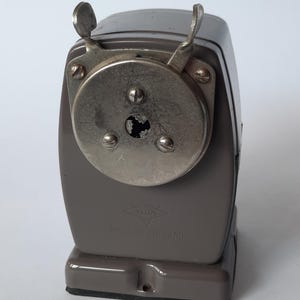 Puede incluir: Un sacapuntas gris vintage con una cubierta de cuchilla y manivela de metal plateado. El sacapuntas tiene una base rectangular y un cuerpo curvo. El texto "VELOR" y "MADE IN HOLLAND" son visibles en la parte delantera.
