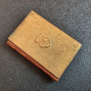 Puede incluir: Un peque&ntilde;o libro de color dorado con un dise&ntilde;o ornamentado y una cresta floral en la portada. El libro est&aacute; encuadernado en cuero marr&oacute;n y tiene un aspecto desgastado y vintage.