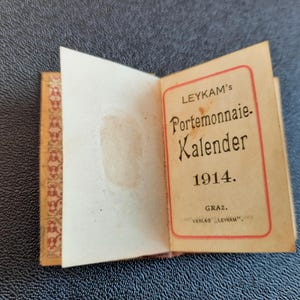 Puede incluir: Un peque&ntilde;o libro vintage marr&oacute;n con una cubierta con estampado dorado y rojo. El libro est&aacute; abierto para revelar una p&aacute;gina de t&iacute;tulo con el texto "LEYKAM's Portemonnaie Kalender 1914. GRAZ. VERLAG "LEYKAM"".