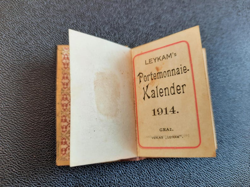 Puede incluir: Un peque&ntilde;o libro vintage marr&oacute;n con una cubierta con estampado dorado y rojo. El libro est&aacute; abierto para revelar una p&aacute;gina de t&iacute;tulo con el texto "LEYKAM's Portemonnaie Kalender 1914. GRAZ. VERLAG "LEYKAM"".