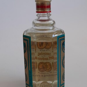 Puede incluir: Una botella de vidrio transparente con una tapa dorada y roja. La botella tiene una etiqueta con texto e ilustraciones de monedas. El l&iacute;quido en el interior es de color amarillo claro. La botella est&aacute; etiquetada como "K&Ouml;LN 187".