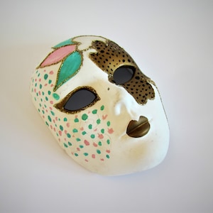 Peut inclure: Un masque de carnaval blanc avec des accents dorés, roses et verts. Le masque a un design décoratif avec des points et des volutes. Il présente une grande ouverture pour les yeux et une petite ouverture pour la bouche.