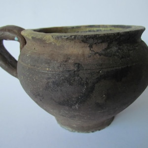 Peut inclure: Un pot en terre cuite brun foncé non verni avec une seule anse. Le pot a un corps rond et un bord légèrement évasé.