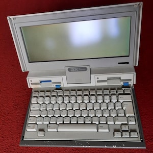 Pode incluir: Um laptop IBM vintage aberto, com teclado e tela. O laptop é cinza claro com um teclado preto e teclas brancas. A tela é cinza escuro. O teclado tem letras e números brancos. O laptop está em uma superfície vermelha.