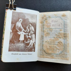 Puede incluir: Libro abierto con ilustraciones en tonos sepia. Una página muestra una imagen religiosa de una mujer y niños, con texto debajo. La página opuesta tiene texto en un marco decorativo. Un clip negro mantiene el libro abierto.