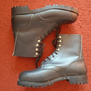 May include: A pair of black leather lace-up boots featuring a high-top design, silver-toned metal eyelets, black laces, and a thick, heavy-duty rubber lug sole with deep treads for traction. The boots have a smooth finish and a rounded toe box.