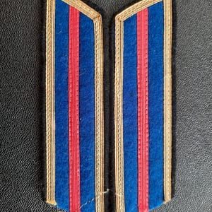 May include: Pair of vintage blue and red striped epaulettes with gold trim. The epaulettes have a rectangular shape with angled edges. The background is a dark, textured surface.