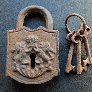 Puede incluir: Un candado y un juego de llaves antiguos y desgastados. El candado es de color marrón óxido, con un emblema decorativo y un ojo de cerradura. Las llaves están unidas a un anillo. El conjunto es probablemente de hierro fundido.
