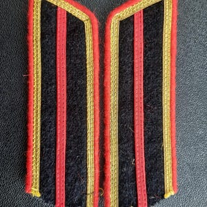May include: Two black fabric shoulder boards with red and gold trim. The boards have two red stripes and a gold border.