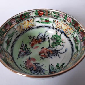 May include: A decorative porcelain bowl with a gold rim and a detailed interior design. The bowl features a central image of stylized birds and plants in shades of red, green, and blue, surrounded by a floral and geometric border.