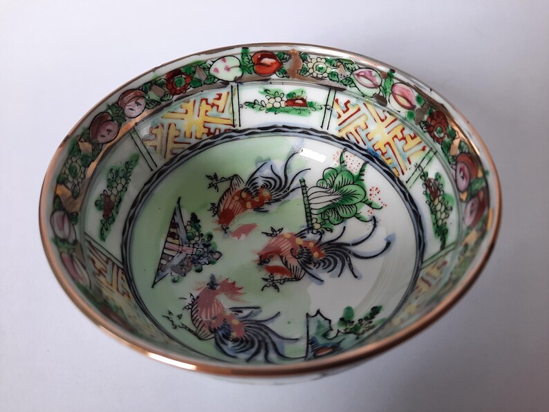 May include: A decorative porcelain bowl with a gold rim and a detailed interior design. The bowl features a central image of stylized birds and plants in shades of red, green, and blue, surrounded by a floral and geometric border.