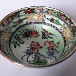 May include: A detailed overhead shot of a decorative porcelain bowl. The bowl features a gold rim and a light green interior with colorful illustrations of birds and figures. The rim is decorated with floral and geometric patterns.