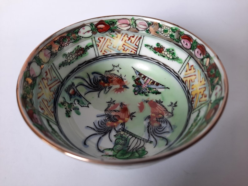 May include: A detailed overhead shot of a decorative porcelain bowl. The bowl features a gold rim and a light green interior with colorful illustrations of birds and figures. The rim is decorated with floral and geometric patterns.