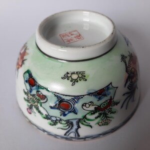 May include: A small, white ceramic bowl with a detailed, hand-painted design. The bowl features a light green background with colorful illustrations of dragons and floral patterns. The rim is white, and the base has a red stamp.