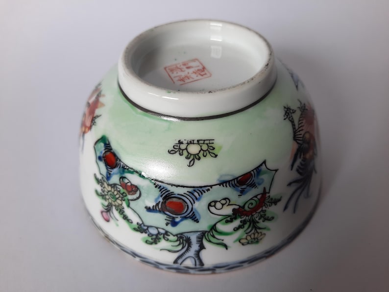 May include: A small, white ceramic bowl with a detailed, hand-painted design. The bowl features a light green background with colorful illustrations of dragons and floral patterns. The rim is white, and the base has a red stamp.
