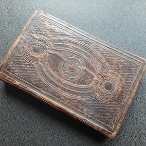May include: A vintage leather-bound book with an intricate embossed design featuring a central circle with a swirling pattern and two smaller circles on either side.