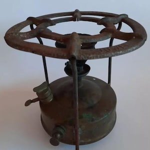 Pode incluir: Um fogão de campismo antigo em latão, com um queimador circular e um depósito de combustível. O fogão tem um aspeto envelhecido, com sinais de idade e uso. O queimador é suportado por finas pernas de metal, e o depósito de combustível tem uma válvula e um botão.