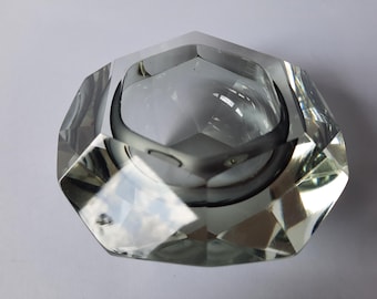 May include: A faceted, clear glass ashtray with a hexagonal shape. The ashtray has a deep, circular bowl in the center and is made of thick, transparent glass. The edges are cut with multiple facets, creating a sparkling effect.