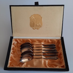 May include: A set of silver spoons in a black box with a gold emblem. The spoons are arranged neatly on a bed of orange fabric. The box is open, revealing the spoons and the emblem.