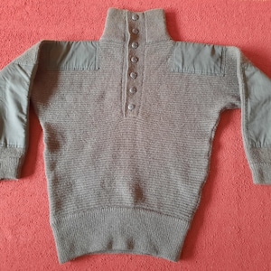 May include: A taupe ribbed knit sweater featuring a high collar with a vertical row of five buttons. The garment includes smooth, solid-colored fabric patches on the shoulders and along the outer sleeves, finished with ribbed cuffs and hem.