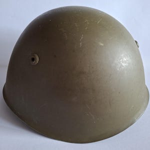 Puede incluir: Un casco de estilo militar de color verde oliva con una forma de cúpula lisa y redondeada. El casco presenta un acabado mate con rozaduras superficiales visibles y pequeños ojales de ventilación circulares en los lados de la carcasa.