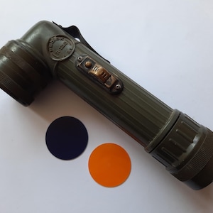 Puede incluir: Una linterna vintage verde oliva con cabezal en ángulo recto, cuerpo acanalado e interruptor de latón. Incluye un filtro de lente circular negro y naranja. La linterna tiene el texto "TL-122/D U.S.A."