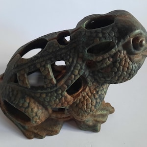 May include: A detailed, weathered frog figurine in shades of green, brown, and bronze. The sculpture has a textured, scaly appearance with open cutouts throughout the body. The frog is posed in a sitting position, showcasing its intricate design.
