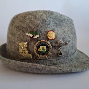 Könnte beinhalten: Ein grauer Filzhut, der mit fünf metallenen Souvenir-Anstecknadeln verziert ist. Die Anstecker zeigen verschiedene Motive, darunter einen Wanderer, ein kreisförmiges Emblem mit einem Baum, eine Seilbahn, ein kleines Porträt und ein Paar Stiefel.