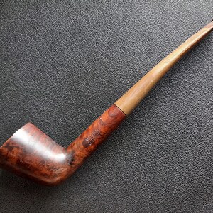 Puede incluir: Una pipa de tabaco con un hornillo de madera marrón oscuro pulido con veta visible y una boquilla larga de madera de color tostado claro. El hornillo tiene grabado el texto VAUEN DE LUXE en una fuente clara y legible en el lateral del caño.