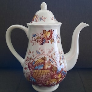 Op de afbeelding: Witte keramische theepot met deksel, handvat en tuit. De theepot is versierd met een kleurrijk fruitmandontwerp, met druiven, perziken en ander fruit. Het deksel en de body hebben bloemaccenten. Een klassiek stuk voor het serveren van thee.