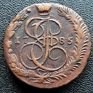Może przedstawiać: Miedziana moneta z monogramem i datą 1785. Moneta posiada koronę i jest otoczona wieńcem laurowym. Powierzchnia monety wykazuje ślady zużycia i wieku.