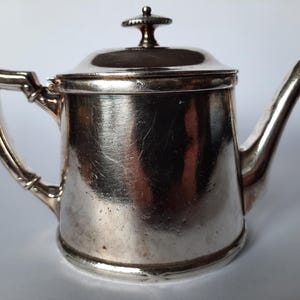 Puede incluir: Pequeña tetera plateada con tapa y boquilla curva. La tetera tiene un asa y un pequeño pomo en la parte superior de la tapa. Probablemente hecha de metal, tiene una superficie reflectante.