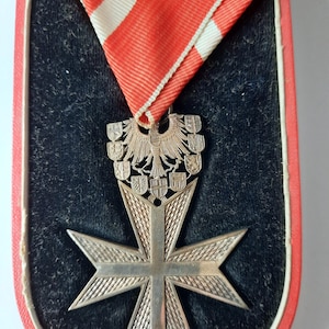 May include: A silver cross with a silver eagle and shields on top, displayed on a black velvet background with a red and white ribbon.