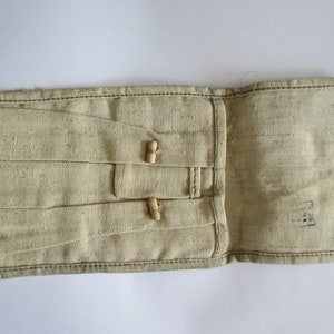 Puede incluir: Una bolsa de lona beige con dos botones de madera y un pequeño sello negro. La bolsa está abierta, revelando el interior.