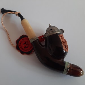 May include: A vintage tobacco pipe with a dark brown bowl and a light-colored stem. The pipe has a metal cap and a carved design on the bowl. A red and orange tag is attached to the pipe.