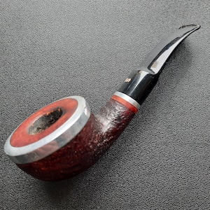 May include: A classic tobacco pipe with a dark brown, textured bowl and a silver rim. The stem is black with a curved, glossy mouthpiece. A red band accents the bowl's base. The pipe rests on a dark surface.