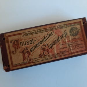 May include: A vintage box of Anusol-Hemorroidalni Suppozitoriji, a hemorrhoid treatment. The box is brown with a label that reads "Suppositoria haemorrhoidalia, Anusol" in white lettering. The label also includes the text "Anusol-Hemorroidalni Suppozitoriji" in a decorative font.